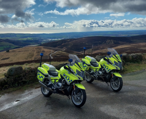 Two police motorcycles in high-visibility yellow parked on a scenic rural hillside road with a vast landscape and cloudy sky in the background.