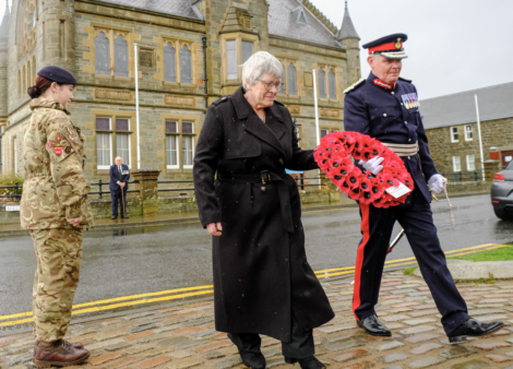 A woman and a man in military uniform hold a wreath of red poppies during a ceremony, with a soldier and a stone building in the background.