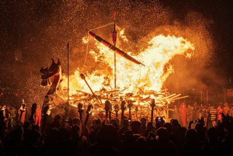 A large wooden structure, resembling a Viking ship, is engulfed in bright flames while an audience watches in the dark, holding torches and capturing the scene with phones.