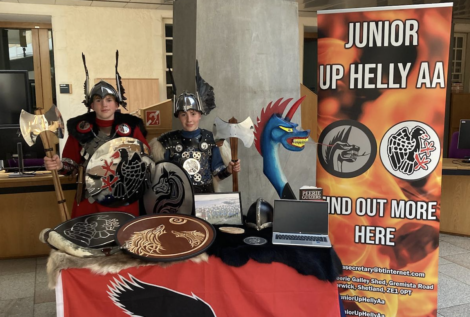 Two children in Viking costumes stand behind a display table with shields, an axe, a dragon head, and a laptop. A banner beside them reads "Junior Up Helly Aa - Find Out More Here.