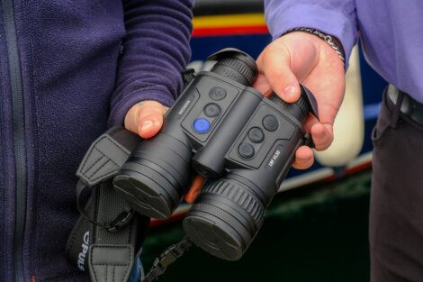 Two individuals hold a pair of black binoculars with various buttons and dials, displaying their features.