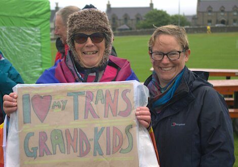 Two people smiling, one holding a sign that reads "I love my trans grand-kids," during an outdoor event. They are dressed in warm clothing and a green field with buildings is visible in the background.