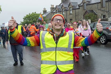 A person wearing a yellow safety vest and rainbow clothing raises both thumbs up while leading a group of people participating in a cheerful parade on a cloudy day.