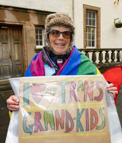 An individual wearing a rainbow flag and a furry hat holds a sign reading "I ❤️ my trans grandkids" outside a building.