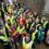 A large group of people in high-visibility vests gather outside a building, looking up at the camera and smiling. Some are dressed in colorful clothing and rain gear.