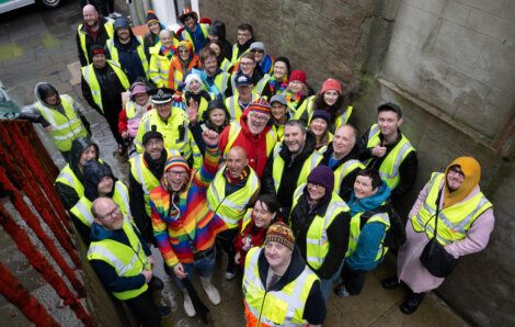 A large group of people in high-visibility vests gather outside a building, looking up at the camera and smiling. Some are dressed in colorful clothing and rain gear.