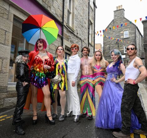 A group of eight people in colorful, flamboyant costumes stand together on a narrow street with stone buildings. One person holds a rainbow umbrella. Bunting is visible in the background.