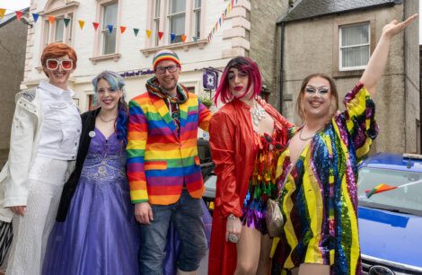 A group of people in colorful and festive outfits pose together on a street. Some wear bright costumes and wigs, and one person is dressed in a rainbow suit. Flags and buildings are in the background.