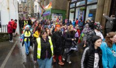 A colorful crowd, some in costumes and high-visibility vests, walks down a wet street during a parade with flags and banners. Buildings and onlookers line the sidewalk.