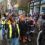 A colorful crowd, some in costumes and high-visibility vests, walks down a wet street during a parade with flags and banners. Buildings and onlookers line the sidewalk.