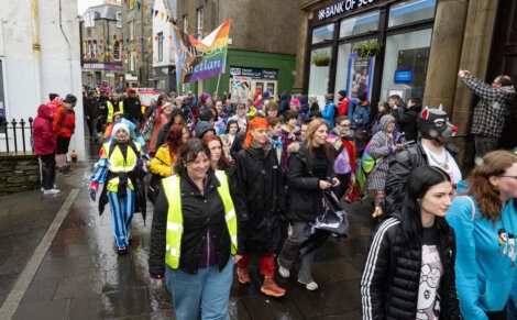 A colorful crowd, some in costumes and high-visibility vests, walks down a wet street during a parade with flags and banners. Buildings and onlookers line the sidewalk.