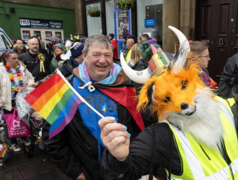 A man smiles while standing next to a person in a fox costume holding a rainbow flag at an outdoor event with other participants in the background.