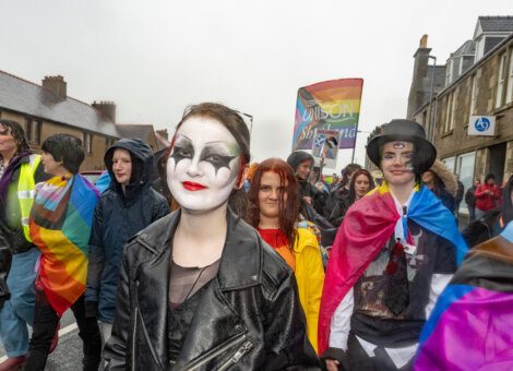 A group of people march in a parade on a rainy day, some wearing colorful outfits and face paint, and carrying rainbow flags.
