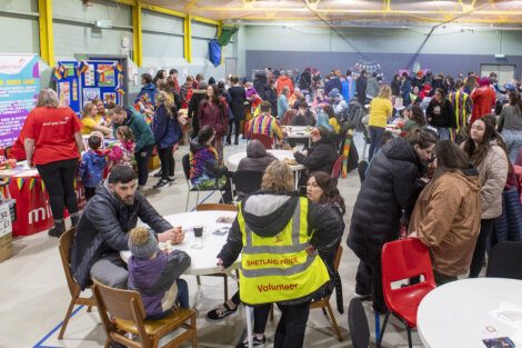 A crowded indoor gathering featuring people of various ages engaging in activities and conversations. A volunteer in a yellow vest stands in the foreground.