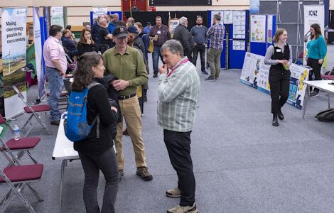 People attending an indoor expo, engaging in conversations with various informational posters and display booths in the background.