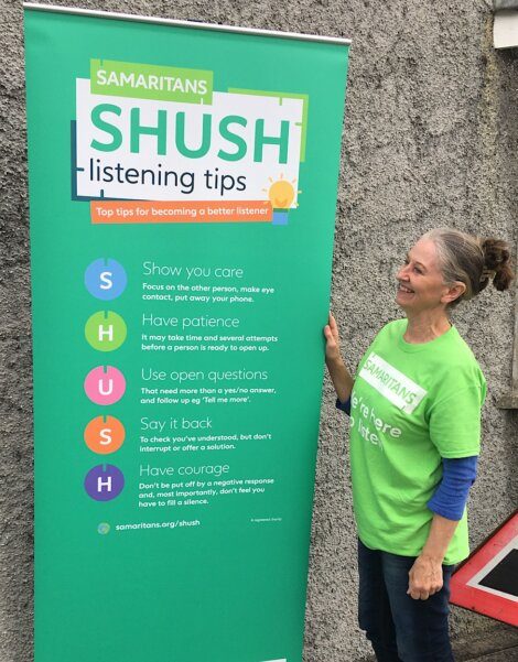A woman wearing a Samaritans t-shirt stands next to a banner displaying SHUSH listening tips: Show you care, Have patience, Use open questions, Say it back, Have courage.