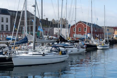 A row of sailboats docked at a marina with buildings in the background under a cloudy sky.