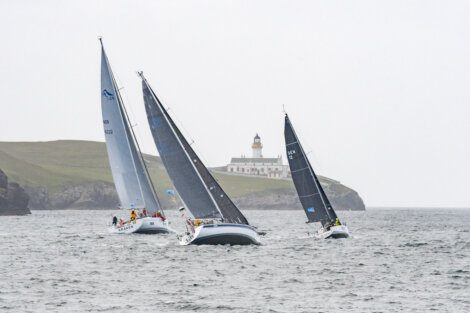 Three sailboats navigate choppy waters near a lighthouse situated on a grassy cliff.