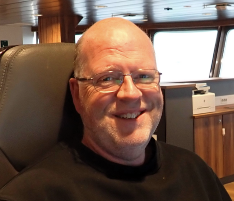 A smiling bald man with glasses and a beard is seated in what appears to be a control room or cockpit. He is wearing a black shirt.