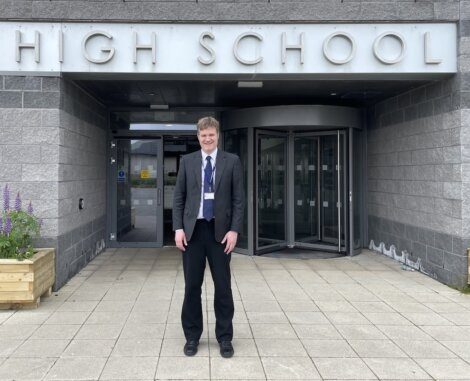 A person stands smiling outside the entrance of a high school building.