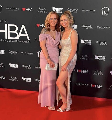 Two women pose together on a red carpet at an event, both dressed formally. One wears a light lavender gown, the other a glittering silver dress with a thigh-high slit. There are event logos in the background.