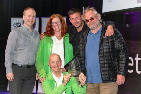 A group of five people standing together and posing for a photo. One person at the front is holding a trophy. They appear to be at an indoor event with banners in the background.