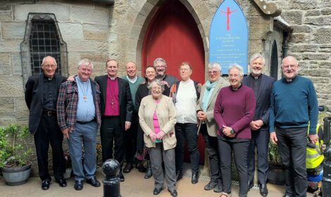A group of people standing and smiling in front of a stone church entrance with a red door and a sign that reads "The Scottish Episcopal Church in Hamilton".