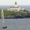 A sailboat with a large white sail is visible in the foreground on the water, with a lighthouse on a rocky hill in the background.