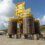 A large yellow offshore subsea structure with four cylindrical legs marked with depth measurements, situated outdoors on a concrete surface against a backdrop of hills and cloudy sky.