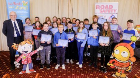 A group of children holding certificates poses with an adult in front of safety-themed banners, including ones for "Go Safe with Ziggy" and "Road Safety." Two cartoon characters are on either side.