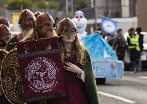 People dressed in Viking costumes walk in a parade for the 2024 Scalloway Fire Festival. One holds a banner with festival details. In the background, a float themed on the character Olaf follows.
