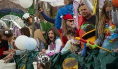 A group of children and adults, some in costumes, are on a parade float decorated with various items. Some hold buckets and balloons. People in the background appear to be also enjoying the event.