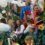 A group of children and adults, some in costumes, are on a parade float decorated with various items. Some hold buckets and balloons. People in the background appear to be also enjoying the event.