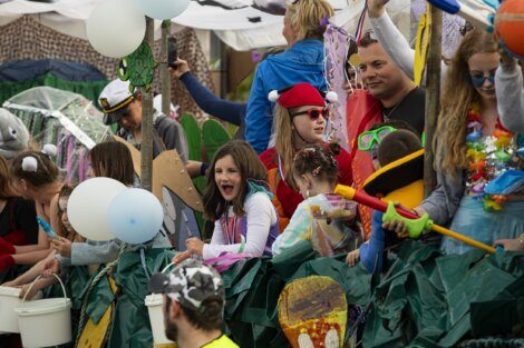 A group of children and adults, some in costumes, are on a parade float decorated with various items. Some hold buckets and balloons. People in the background appear to be also enjoying the event.