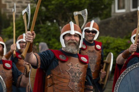 A group of people dressed as Norse warriors with helmets, shields, and axes participate in a historical reenactment outdoors.