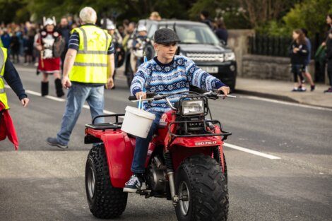 A boy wearing a patterned sweater and a flat cap rides a red ATV carrying a white bucket during a parade. Other participants in reflective vests and costumes are visible in the background.
