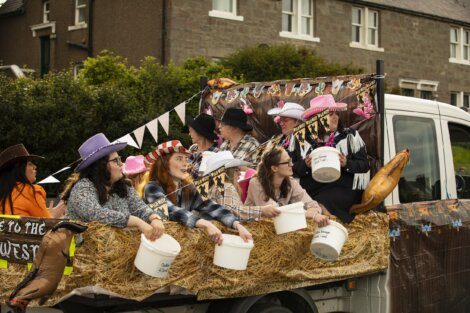 A group of people dressed in cowboy hats ride on a truck decorated with hay bales, holding buckets, in a parade.