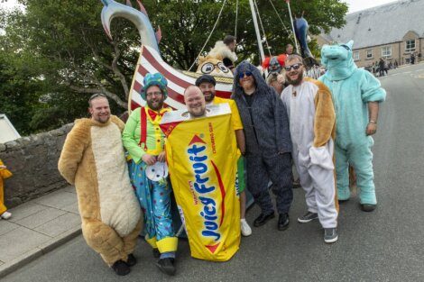 A group of people in various costumes, including a Juicy Fruit wrapper and animal suits, stand together on a street with a Viking ship in the background.