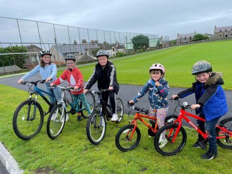 Five children in helmets stand with their bicycles on a paved path next to a grassy field and fence, with houses visible in the background on a cloudy day.