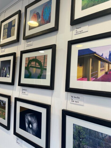 A gallery wall displaying multiple framed photographs with labels underneath each. One prominent photo depicts a wreath; another features a pavilion.