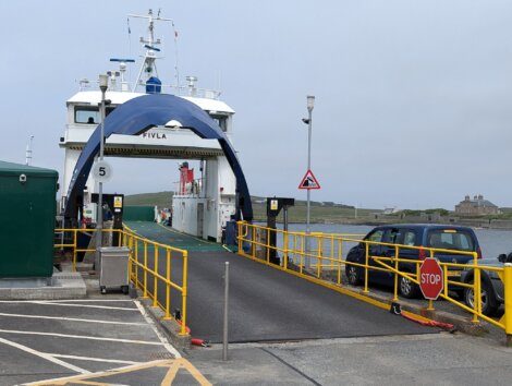 Ferry docked at port with ramp extended, surrounded by yellow railings and traffic signs. Cars and a building are visible in the background.