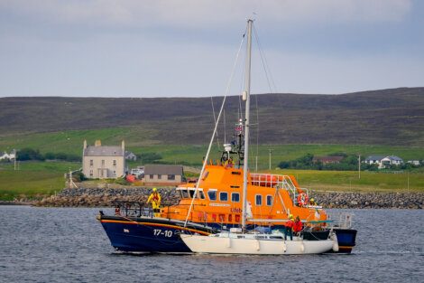 An orange lifeboat assists a white sailboat near a coastal shoreline with houses in the background.