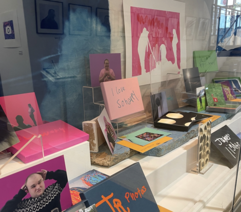 An art display featuring various colorful prints, books, and postcards arranged on shelves and stands, seen through a reflective glass.