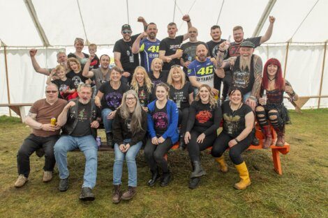 A group of people, men and women, of various ages, some with children, pose enthusiastically inside a tent. They are casually dressed, and a few wear graphic tees and biker gear.