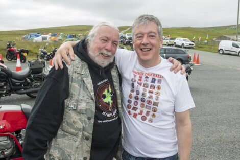 Two men standing and smiling outdoors, with motorcycles and camping tents in the background. One man is wearing a black hoodie and camo vest, while the other is wearing a white t-shirt.