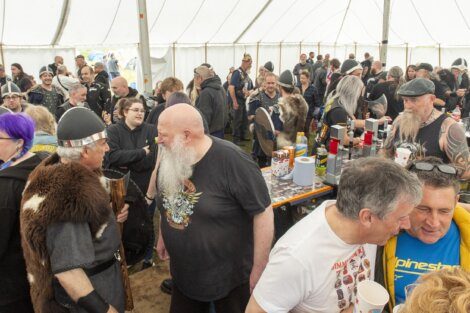 A large group of people, many in casual attire and some in Viking-themed costumes, interact inside a big tent during a daytime event. Several are holding cups and engaging in conversation.