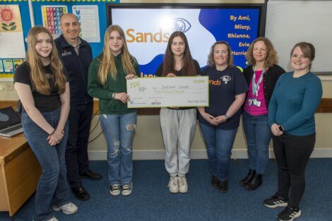 A group of six young women and a man hold a large check for £3,000 made out to Shetland Sands in a classroom setting. The wall behind them features a presentation screen displaying "Sands.