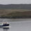 A small ship sails through calm waters with hills and a few buildings visible in the background.