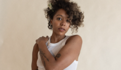 A person with curly hair and tattooed arms wearing a white sleeveless shirt, looking directly at the camera.