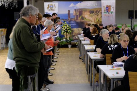 People are gathered in a room, some seated and some standing, engaged in counting activities. A backdrop displays images of the Orkney Islands with "Orkney" written at the top.